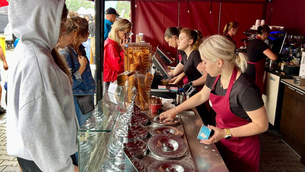 Stoisko lodziarskie Cukierni Sowa z obsługą w bordowych fartuchach, klientami w kolejce i bogatą ofertą lodów w metalowych pojemnikach