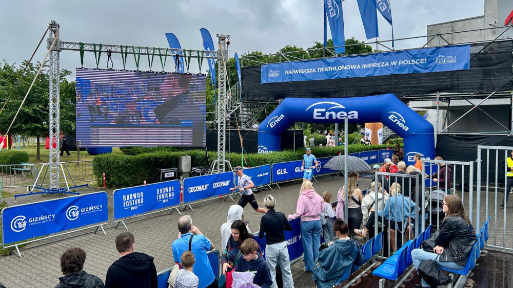 Strefa mety największej imprezy triathlonowej w Polsce z widocznym ekranem LED, balonem Enea oraz tłumem kibiców na trybunach i wzdłuż trasy