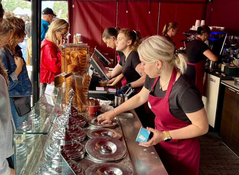 Stoisko lodziarskie Cukierni Sowa z obsługą w bordowych fartuchach, klientami w kolejce i bogatą ofertą lodów w metalowych pojemnikach