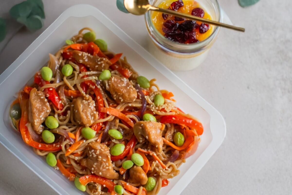 Danie z makaronem, kurczakiem i warzywami posypane sezamem w pojemniku, obok deser w słoiczku z musem owocowym i suszoną żurawiną, na stole udekorowanym gałązkami eukaliptusa.